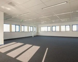 Lichte, lege kantoorruimte met blauw tapijt en grote ramen aan de Capronilaan in Schiphol-Rijk.