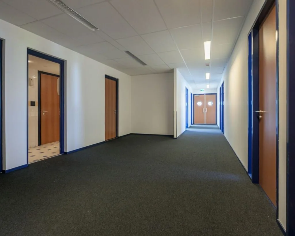 Lange, smalle gang in een kantoorruimte aan de Capronilaan in Schiphol-Rijk met meerdere houten deuren en witblauwe wanden.