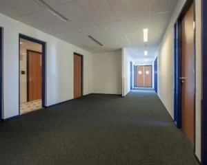 Lange, smalle gang in een kantoorruimte aan de Capronilaan in Schiphol-Rijk met meerdere houten deuren en witblauwe wanden.
