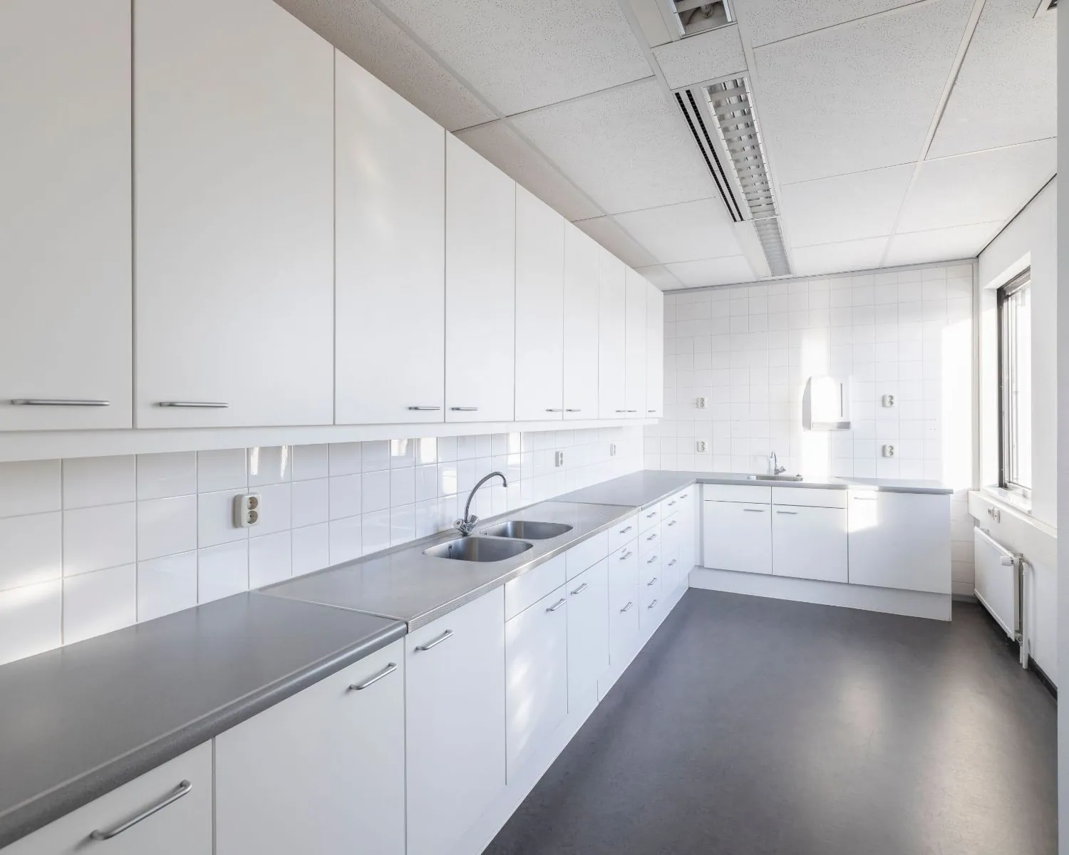 Moderne, lichte kantoorkeuken met witte kasten, dubbele spoelbak en grijs werkblad aan de Stephensonweg 6-8.