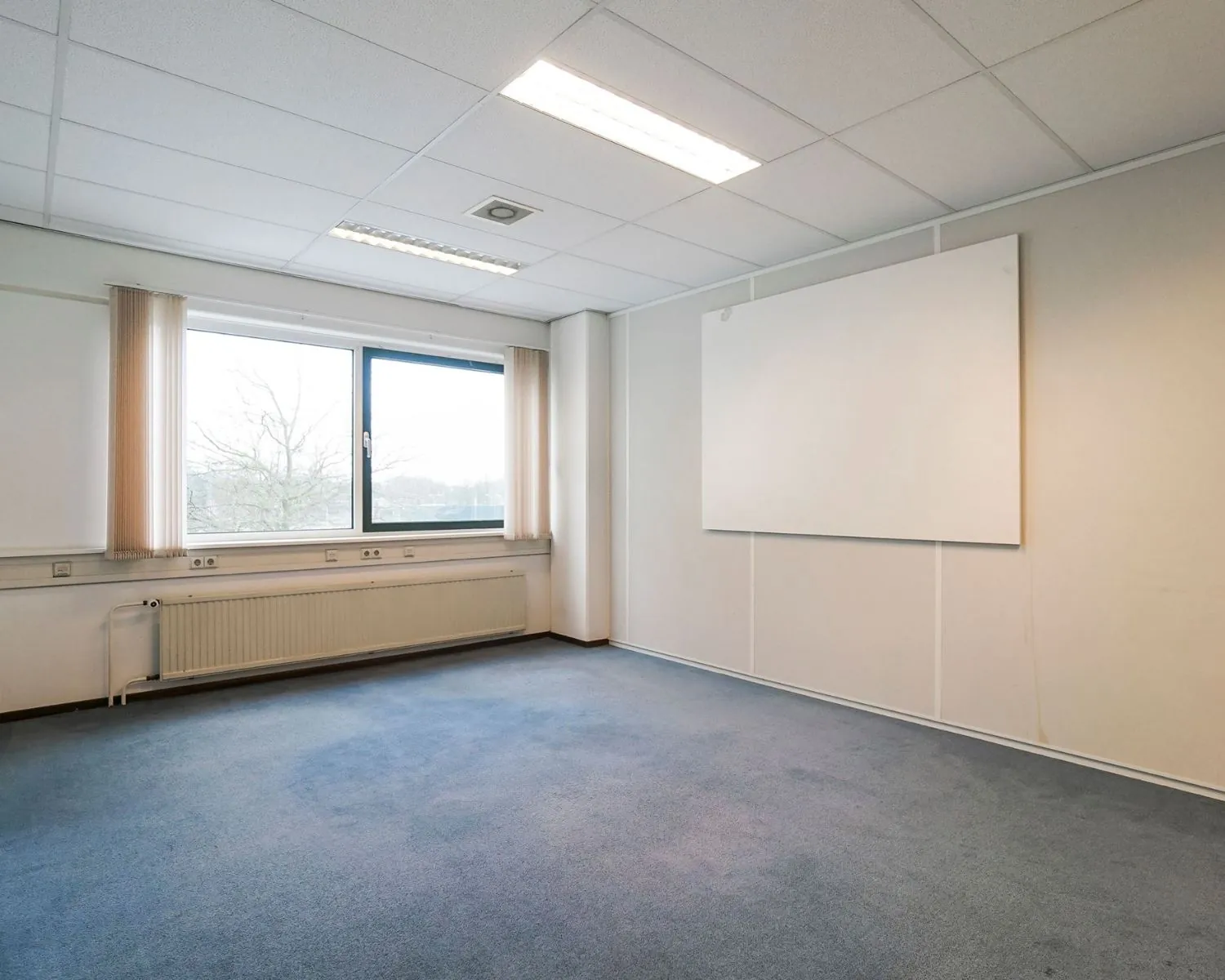 Lege kantoorruimte met blauw tapijt, groot raam en whiteboard aan de muur aan de Haagse Schouwweg 8.