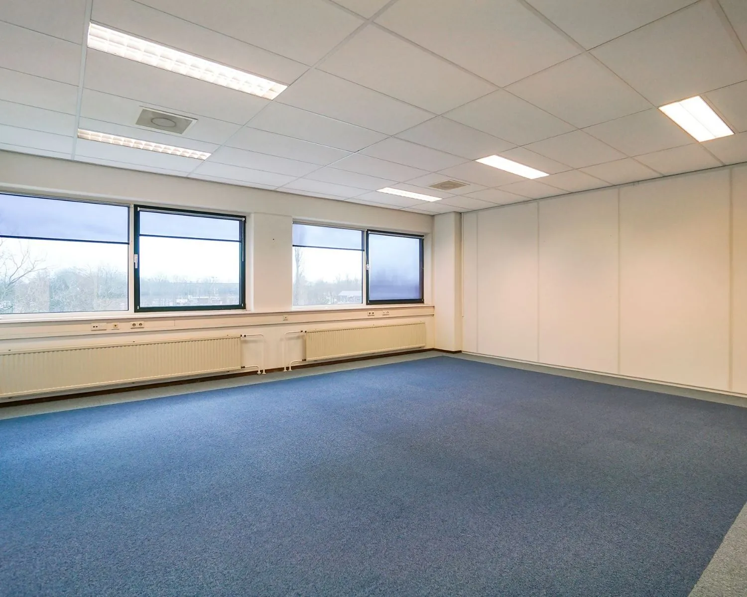 Lichte, lege kantoorruimte met blauwe vloerbedekking aan de Haagse Schouwweg 8.
