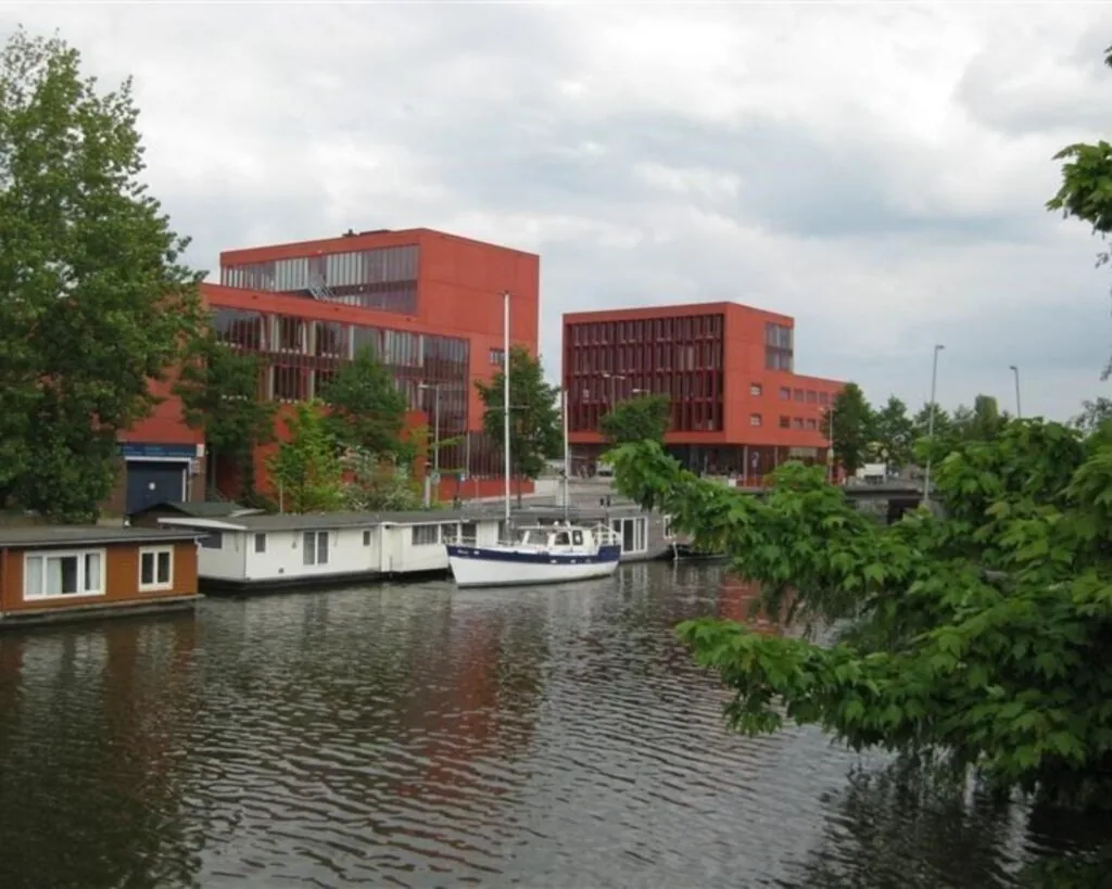 Twee moderne rode kantoorgebouwen aan de Panamalaan in Amsterdam, gelegen aan het water met woonboten en een zeilboot op de voorgrond.