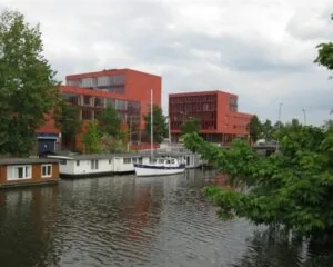 Twee moderne rode kantoorgebouwen aan de Panamalaan in Amsterdam, gelegen aan het water met woonboten en een zeilboot op de voorgrond.