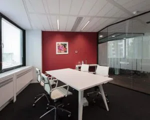 Moderne kantoorruimte met witte bureaus, glazen wand en een rode accentmuur aan de Saturnusstraat 46 in Hoofddorp.
