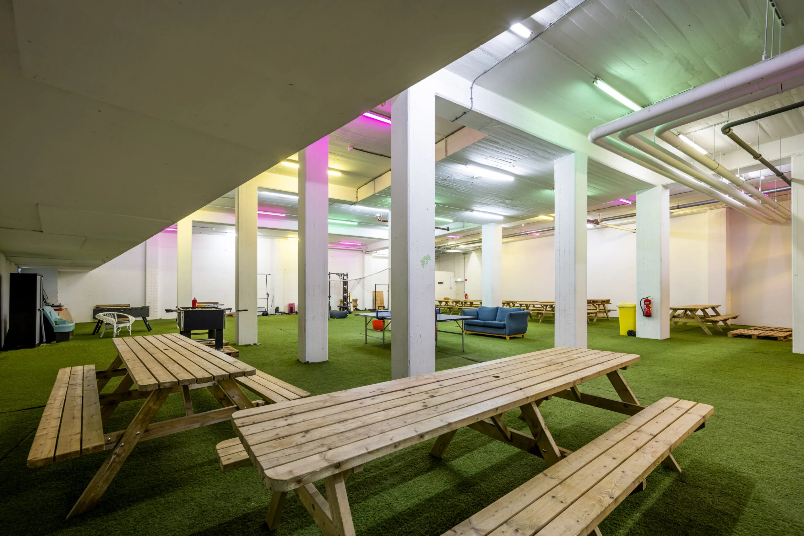 Ruime kelderruimte aan de Singel 542 in Amsterdam met picknicktafels, kunstgrasvloer en kleurrijke verlichting, ingericht als ontspannings- en werkplek.
