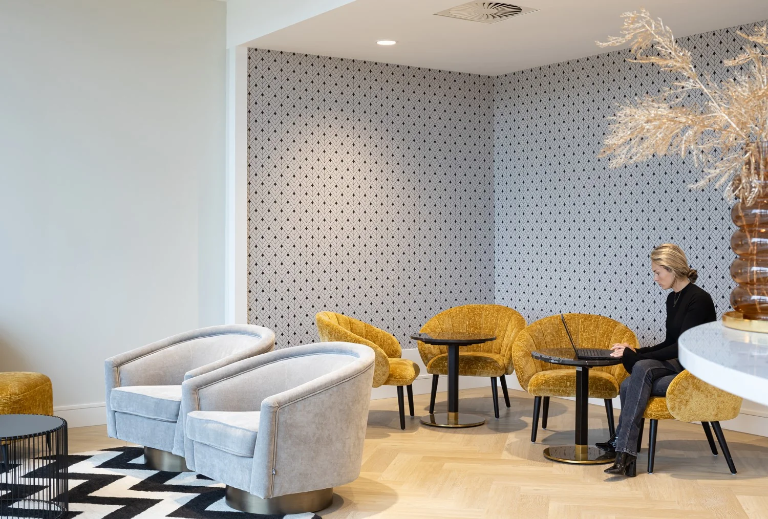 Moderne loungehoek in een kantoorruimte in Hoofddorp met gele en grijze fauteuils, waar een vrouw op een laptop werkt.