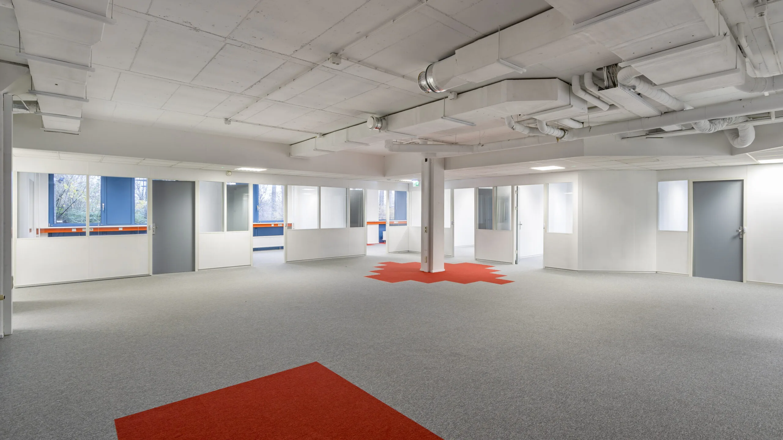 Moderne, lichte kantoorruimte aan de Innsbruckweg 103 in Rotterdam met grijze vloerbedekking, rode accenten en glazen scheidingswanden.