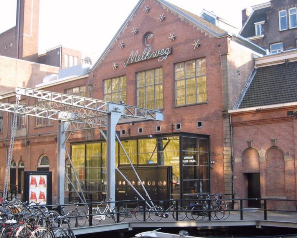 Vooraanzicht van het historische Melkweg-gebouw aan de Korte Leidsedwarsstraat 12 in Amsterdam, met fietsenrekken op de voorgrond.