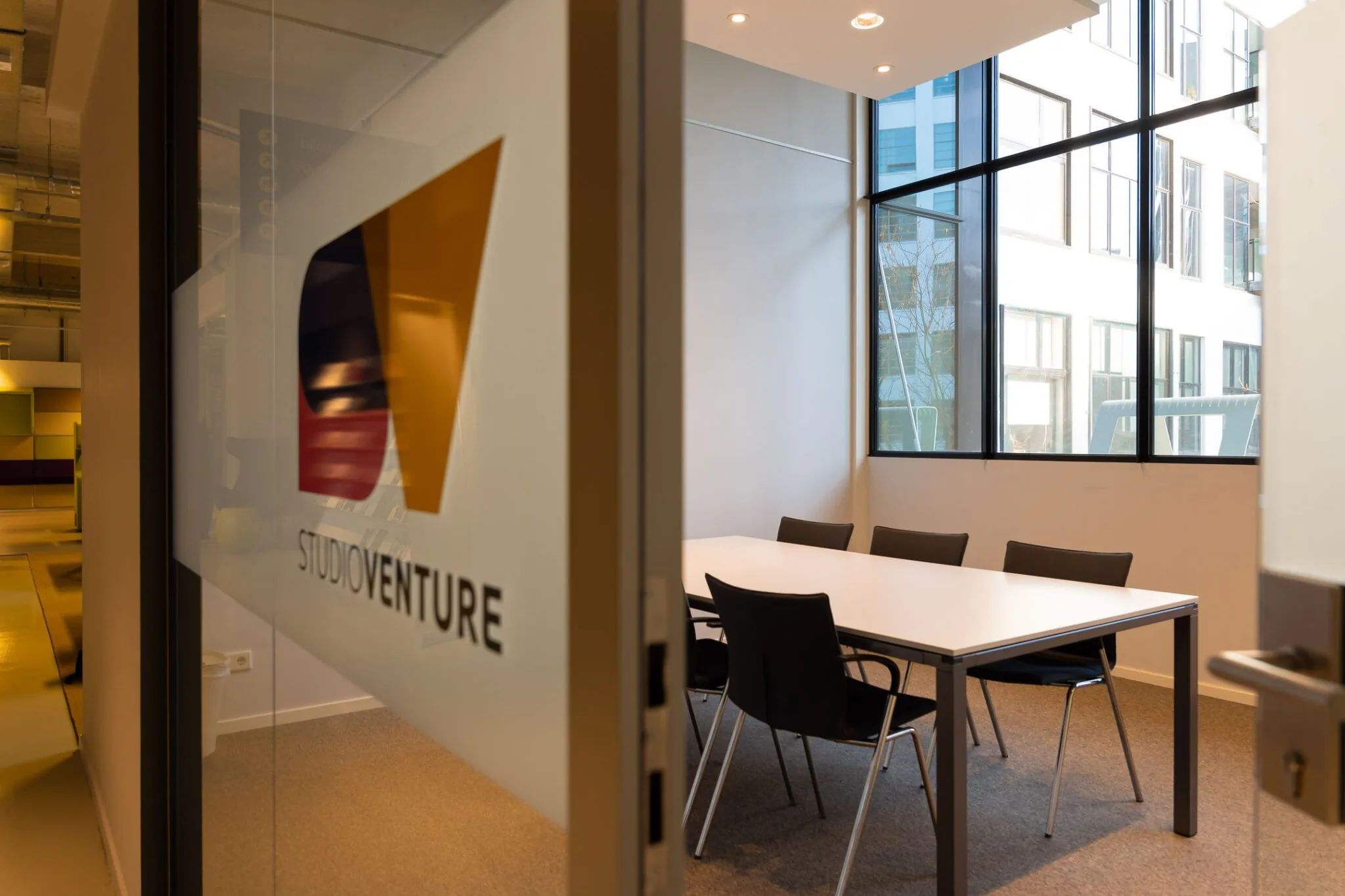 Vergaderruimte met zes stoelen in de Lichttoren 32 in Eindhoven, gezien door een glazen deur met het logo van STUDIOVENTURE.