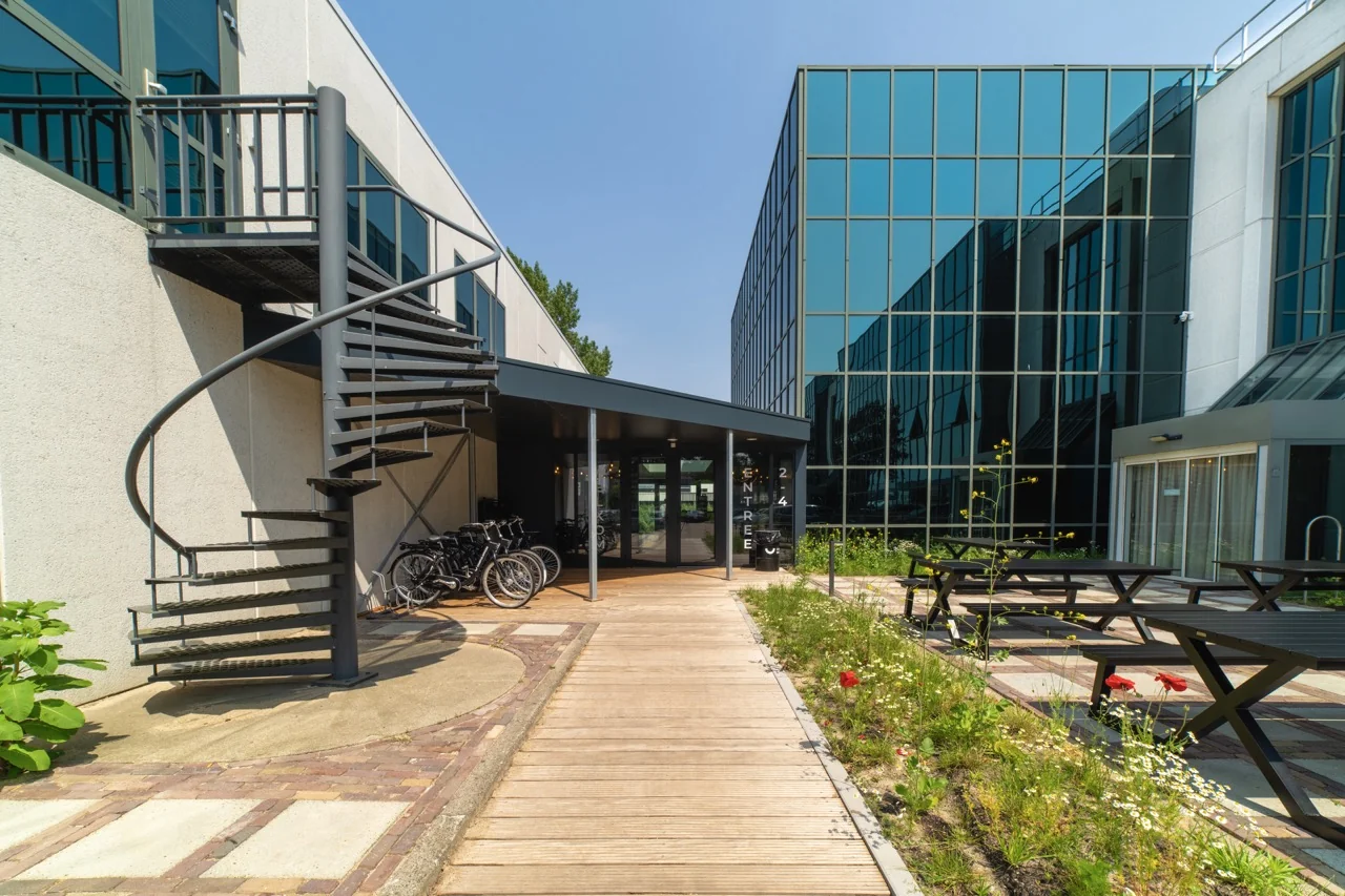 Ingang van een modern kantoorgebouw aan de Rietbaan 2-4-12 in Capelle aan den IJssel met glazen gevel, buitentrap en picknicktafels.