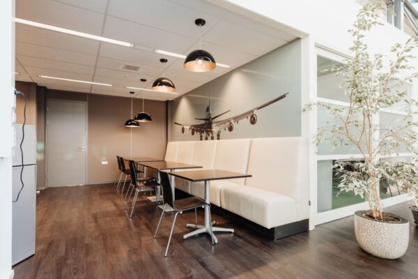Moderne kantine met zitplaatsen, houten vloer, hanglampen en een wanddecoratie van een landend vliegtuig in kantoorruimte aan Sloterweg 303 in Badhoevedorp.