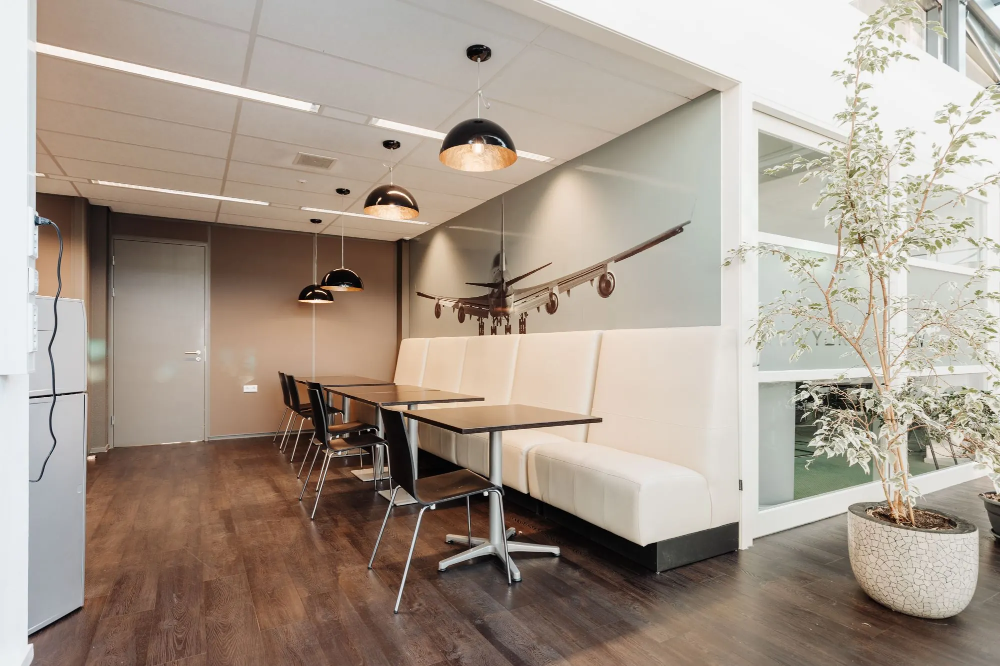 Moderne kantine met zitplaatsen, houten vloer, hanglampen en een wanddecoratie van een landend vliegtuig in kantoorruimte aan Sloterweg 303 in Badhoevedorp.