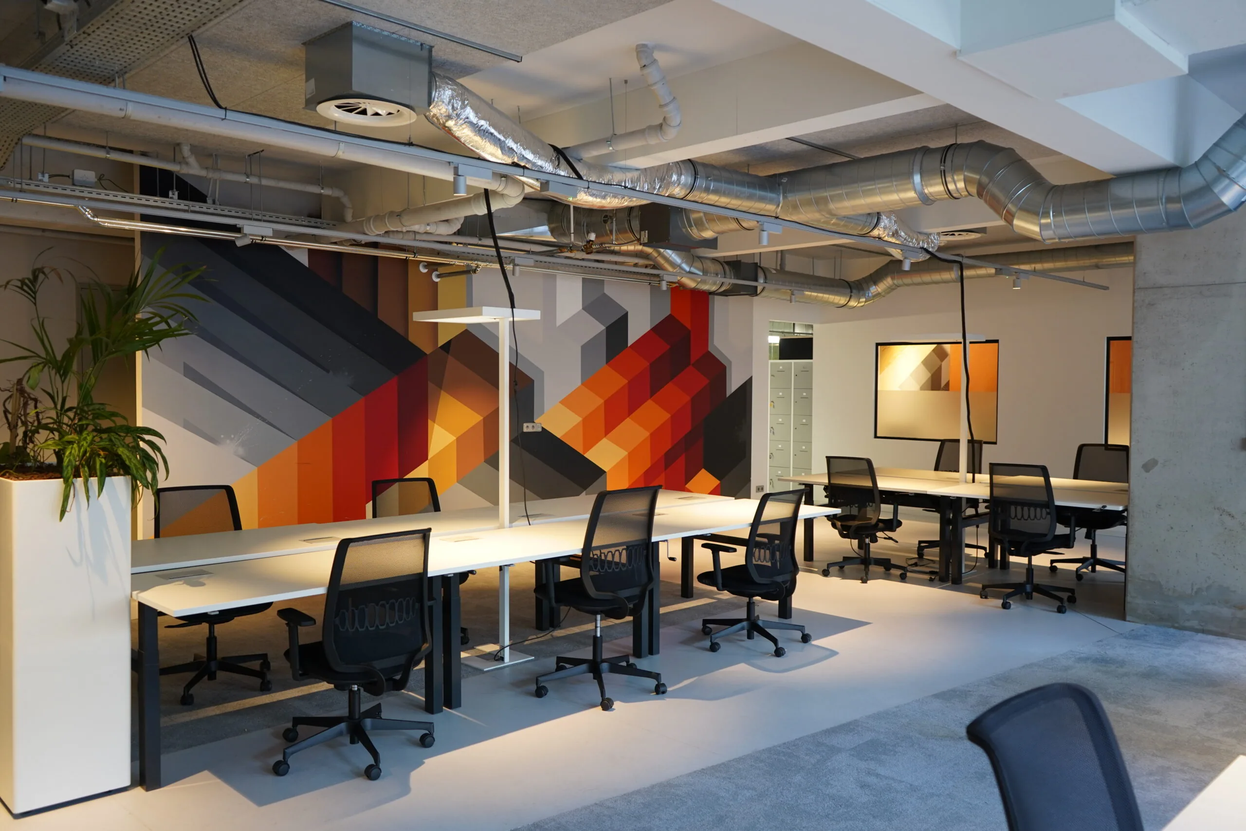 Moderne open kantoorruimte aan het Weesperplein 4A in Amsterdam met bureaus, bureaustoelen en een kleurrijke geometrische muurschildering.