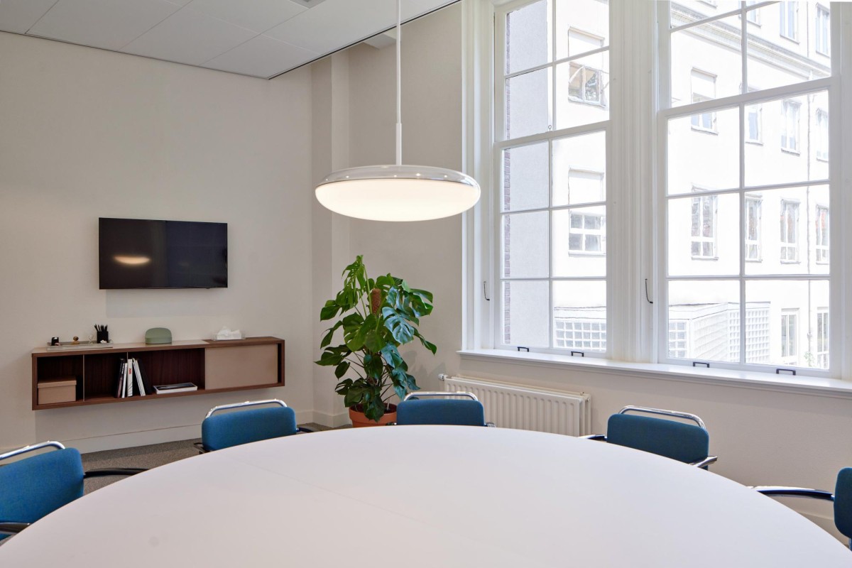 Een vergaderruimte met een ronde tafel, blauwe stoelen, grote ramen en een hanglamp aan de Keizersgracht 555 in Amsterdam.