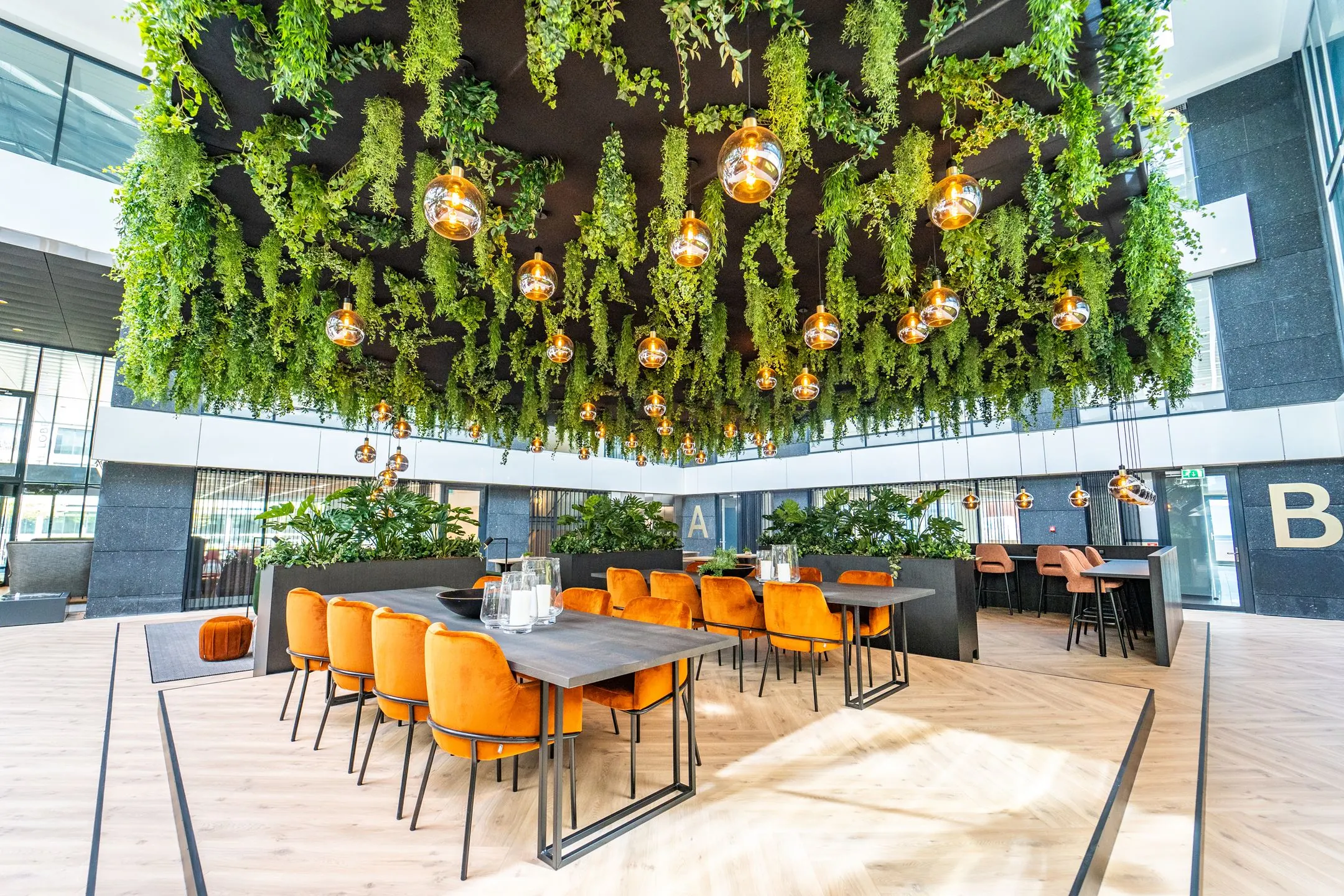 Moderne kantoorruimte aan de Lange Kleiweg met oranje stoelen, houten tafels en een plafond met hangende planten en sfeerverlichting.