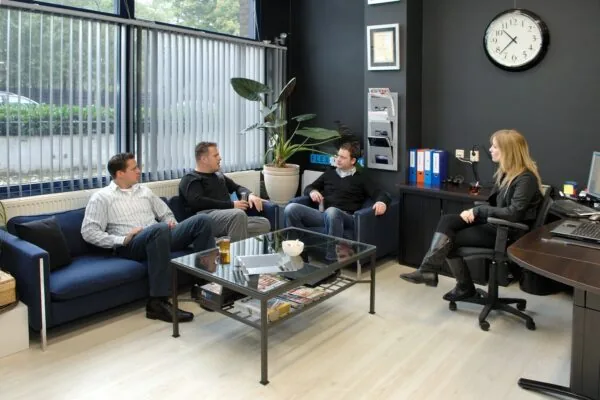 Vier mensen zitten in een moderne kantoorruimte aan de Kanaaldijk-Zuid 19 in gesprek met een vrouw achter een bureau.