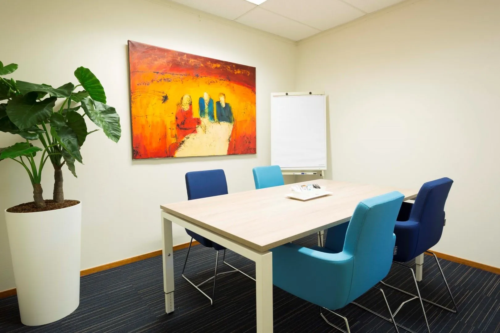 Vergaderruimte aan de Steenovenweg 5 met moderne meubels, kleurrijk schilderij, flip-over en grote kamerplant.