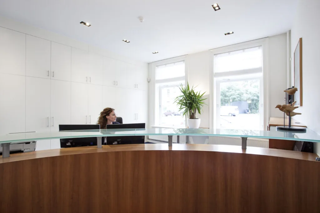 Moderne ontvangstruimte met houten balie, glazen blad en een receptioniste bij kantoorruimte Sweelinckplein 1-3.