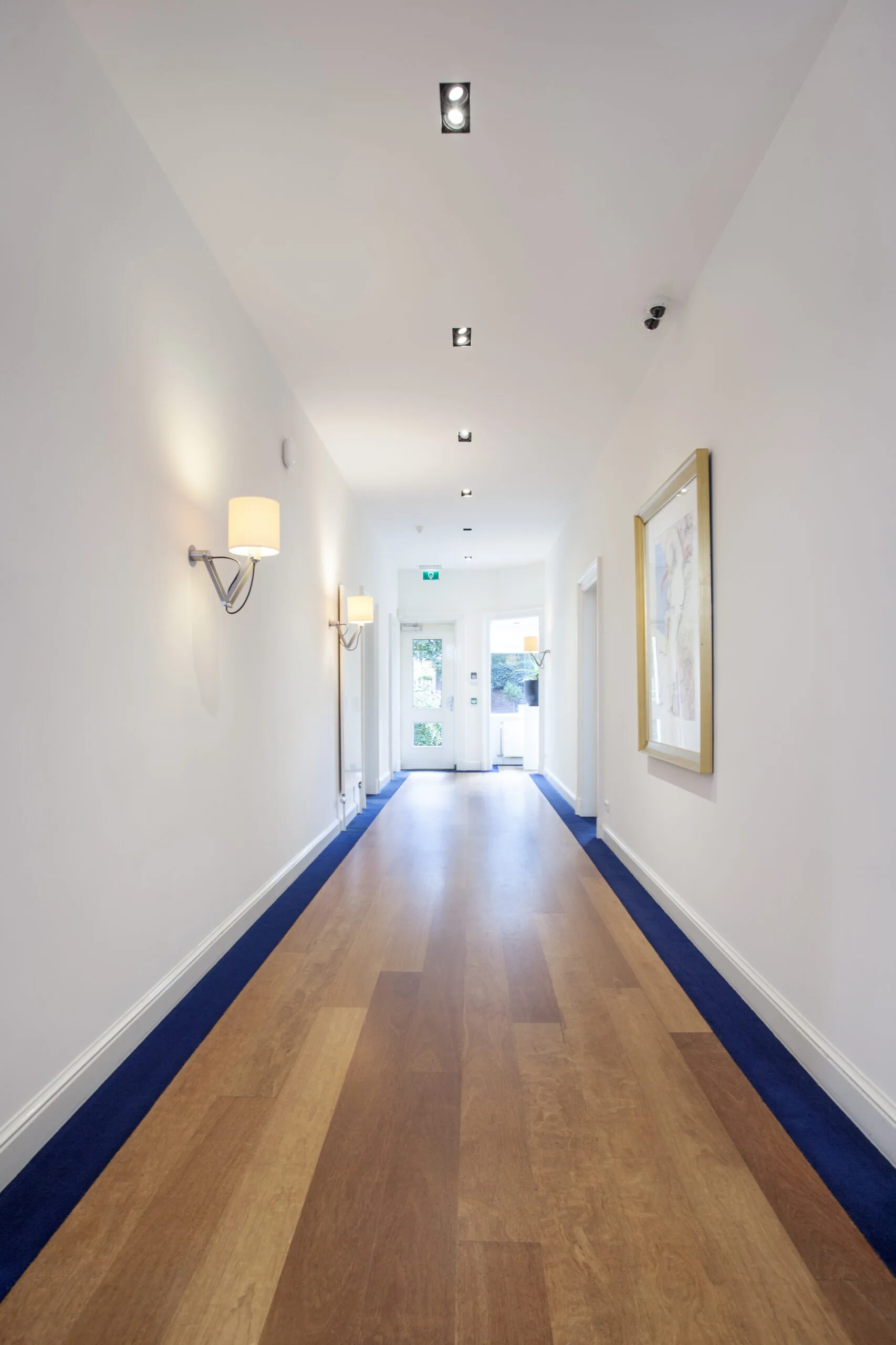 Verlichte gang met houten vloer, witte muren en moderne wandlampen in kantoorruimte Sweelinckplein 1-6.