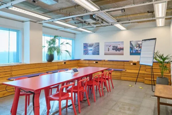 Vergaderruimte met rode tafel en stoelen, houten zitbanken, een flip-over en wandfoto’s aan de Johan Huizingalaan 400.