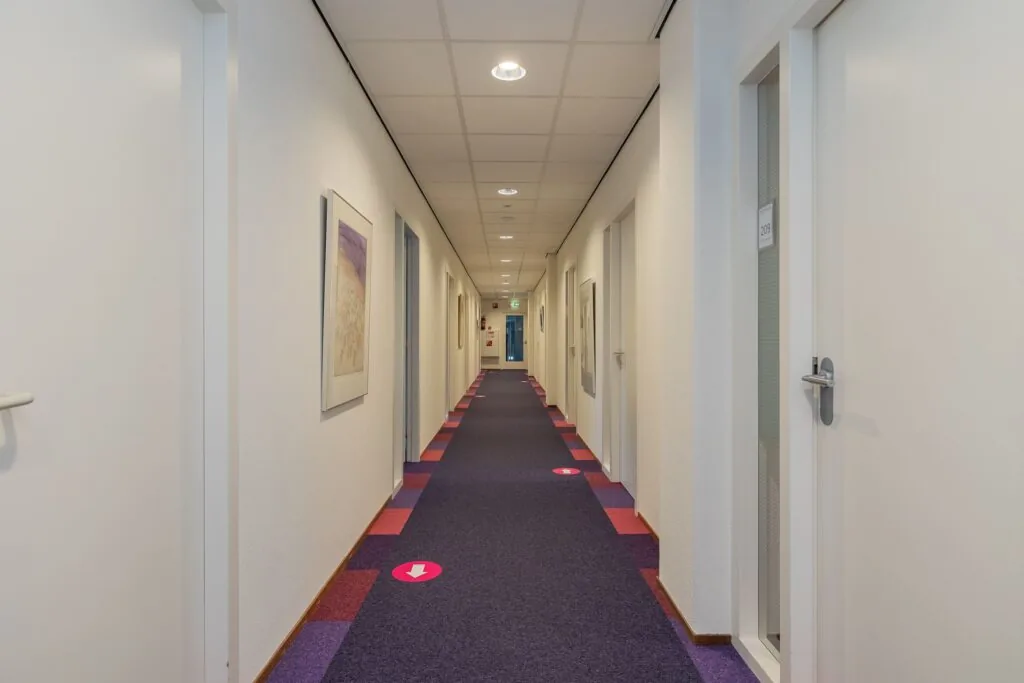 Lange, rechte gang in een kantoorruimte aan de Schipholweg 55 in Leiden met kleurrijk tapijt, witte deuren en kunst aan de muren.