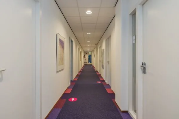 Lange, rechte gang in een kantoorruimte aan de Schipholweg 55 in Leiden met kleurrijk tapijt, witte deuren en kunst aan de muren.