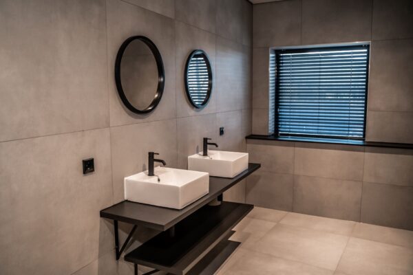 Moderne, minimalistische badkamer in kantoorruimte Reitscheweg 1-8 met twee wasbakken, ronde spiegels en grijze wandtegels.