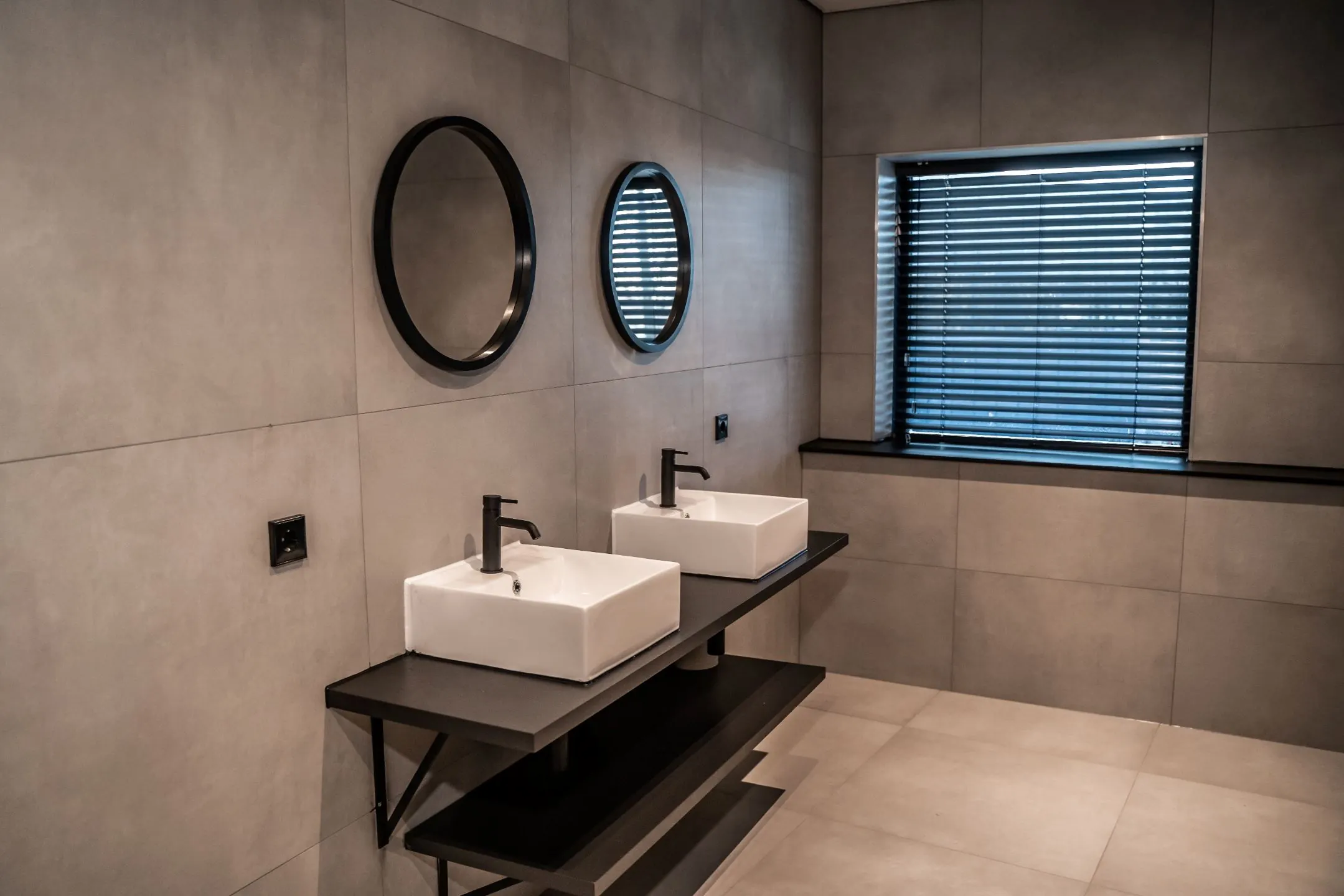 Moderne, minimalistische badkamer in kantoorruimte Reitscheweg 1-8 met twee wasbakken, ronde spiegels en grijze wandtegels.