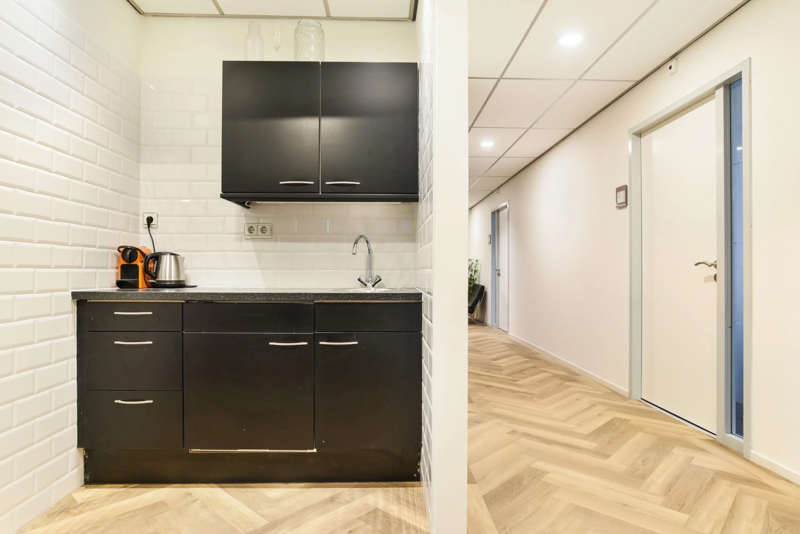 Kleine pantry met zwarte keukenkastjes en koffiezetapparaat naast een gang in een kantoorpand aan het Kennemerplein 20 in Haarlem.