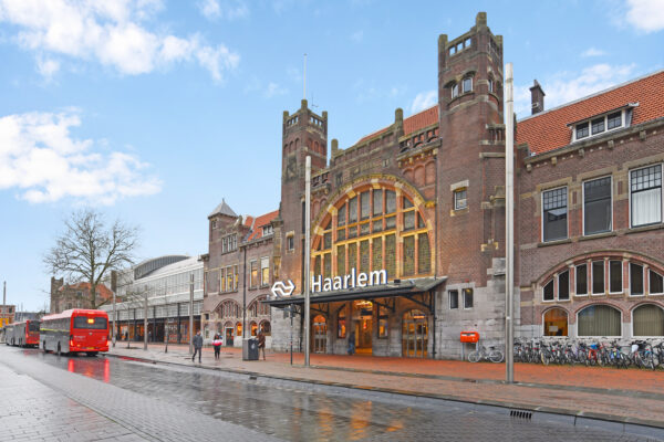 Voorgevel van station Haarlem aan het Kennemerplein met wachtende bussen, fietsen en passerende voetgangers.