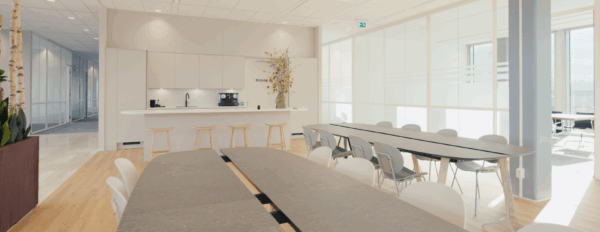 Moderne kantoorkeuken met strakke witte kasten, barkrukken en lange vergadertafels in een lichte ruimte.