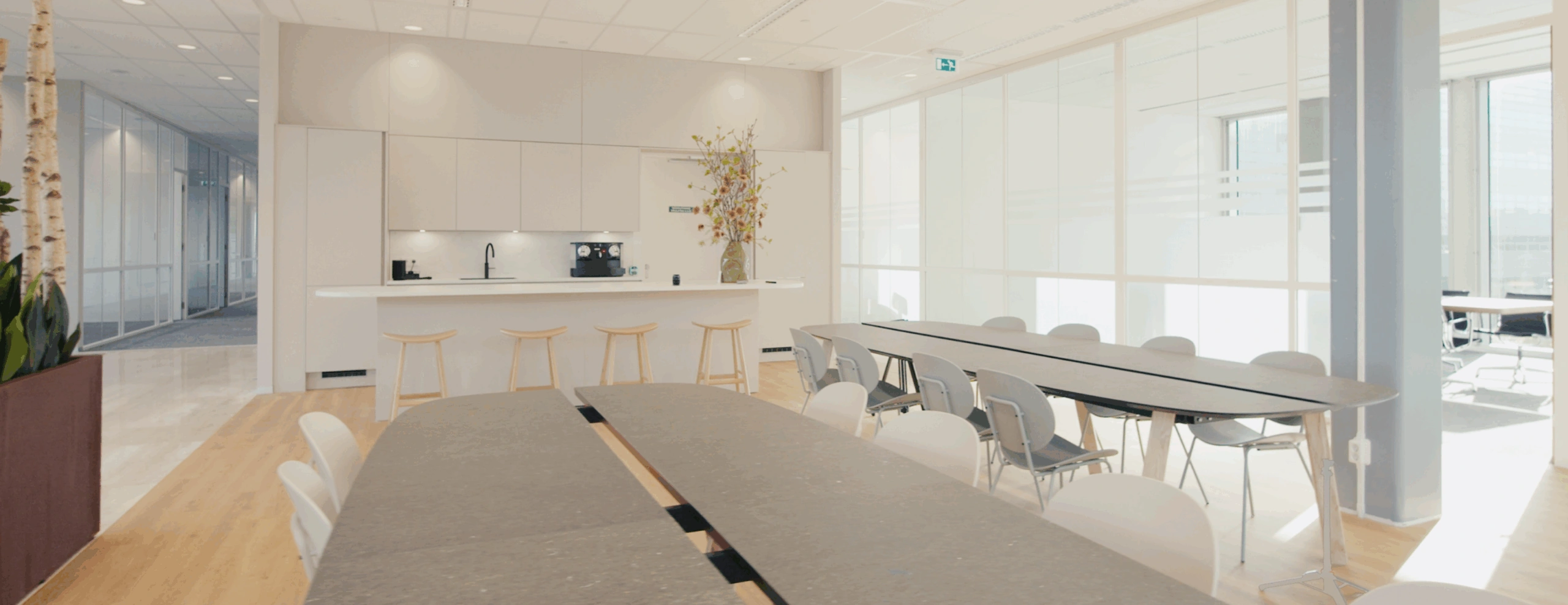 Moderne kantoorkeuken met strakke witte kasten, barkrukken en lange vergadertafels in een lichte ruimte.