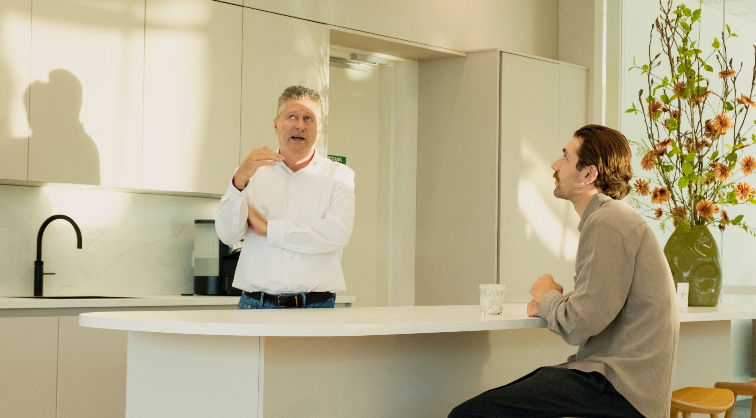 Twee mannen in gesprek in een moderne, lichte keuken met een wit kookeiland en een groot bloemstuk.
