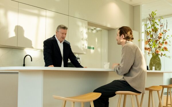 Twee mannen voeren een gesprek aan een keukeneiland in een moderne, licht ingerichte keuken met een grote vaas bloemen op de achtergrond.