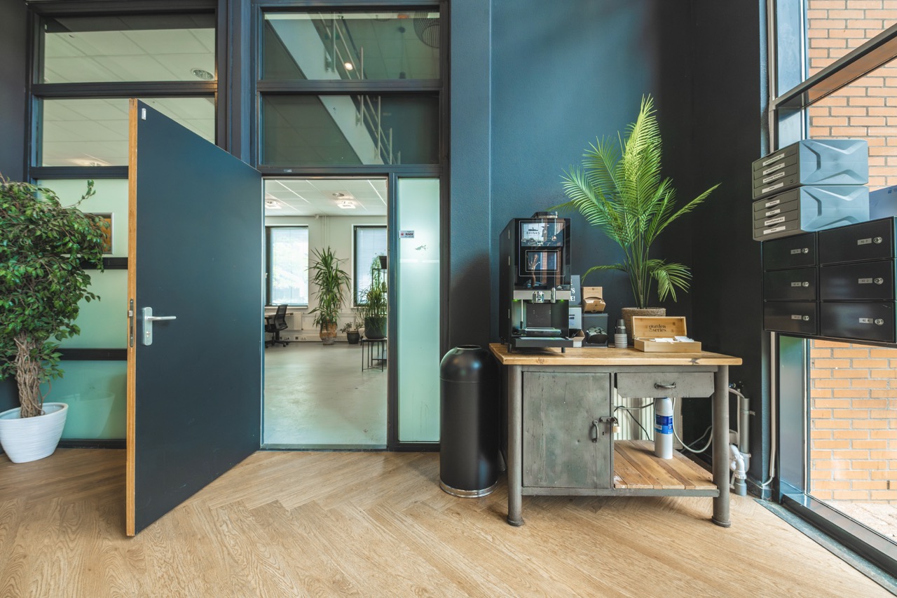 Groot koffiezetapparaat op een metalen tafel in een moderne kantooromgeving met planten en postvakken.