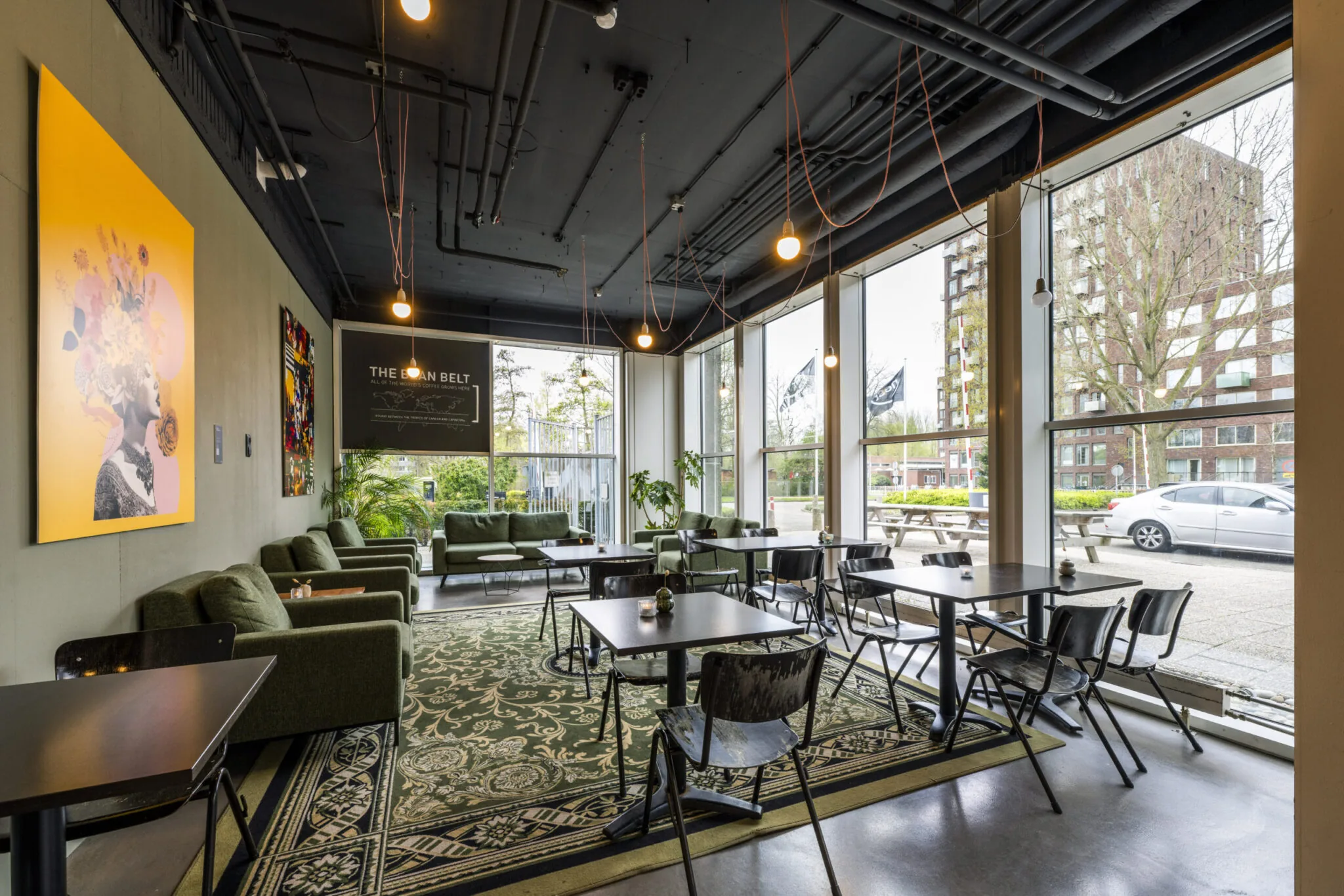 Modern ingerichte lounge- en werkruimte met grote ramen, groene banken, kunst aan de muur en uitzicht op een stedelijke omgeving.