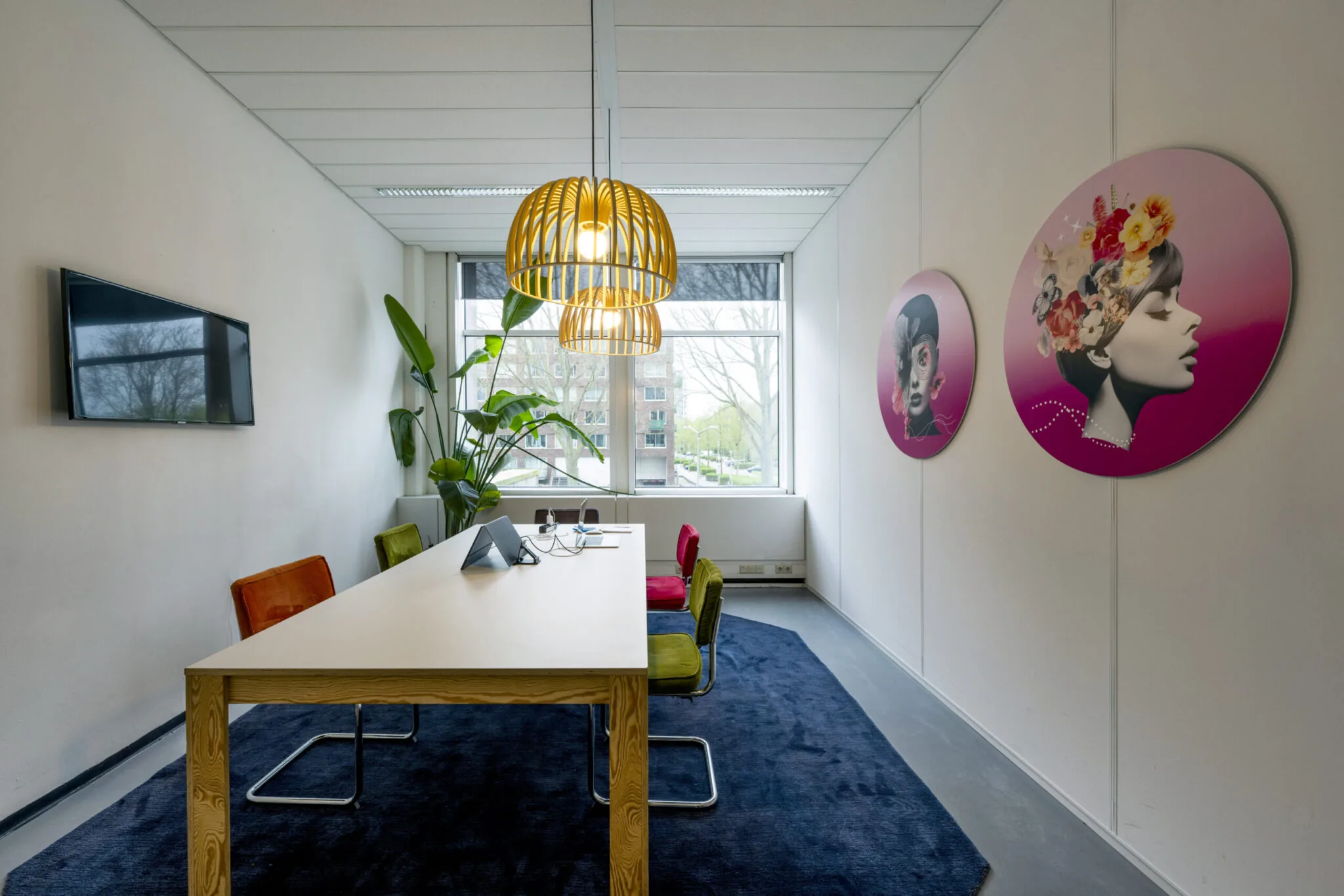 Vergaderruimte met kleurrijke stoelen, houten tafel, grote plant, moderne kunst aan de muur en uitzicht op gebouwen door het raam.