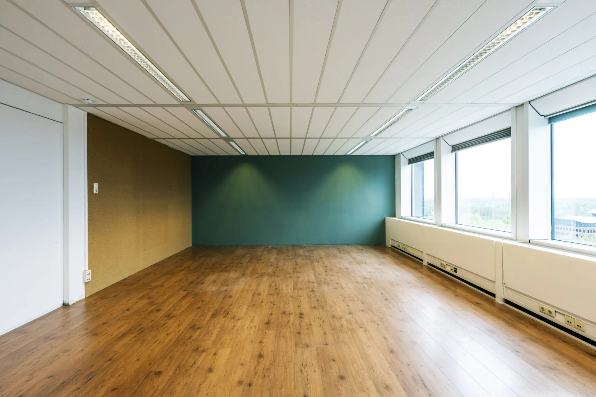 Leeg kantoorruimte in Kronenburg 033 met houten vloer, groenblauwe en kurkwand en grote ramen met uitzicht.