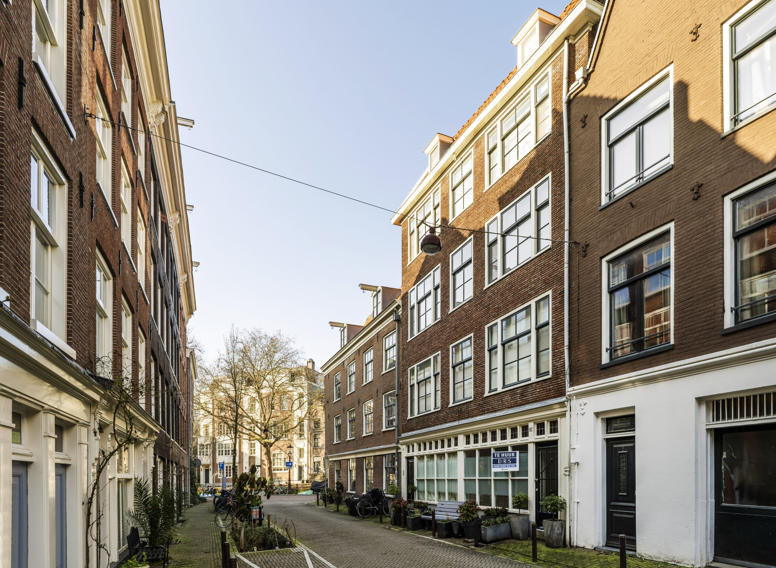 Straatbeeld van de Leliestraat in Amsterdam met karakteristieke bakstenen woningen en een "Te Huur" bord voor een bedrijfsruimte.