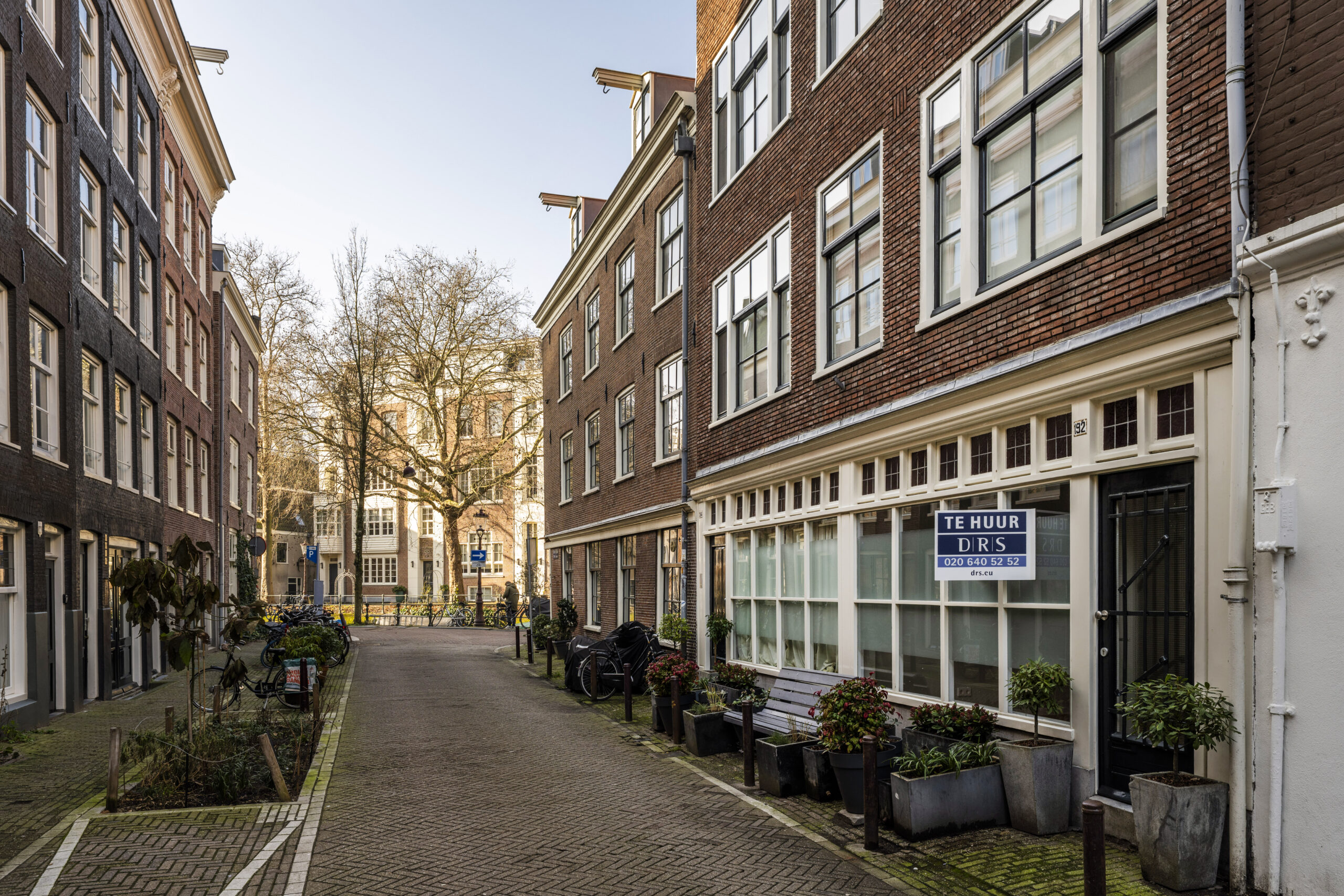 Een rustige straat met klassieke bakstenen panden aan de Leliestraat in Amsterdam, met een te huur-bord op een van de gebouwen.