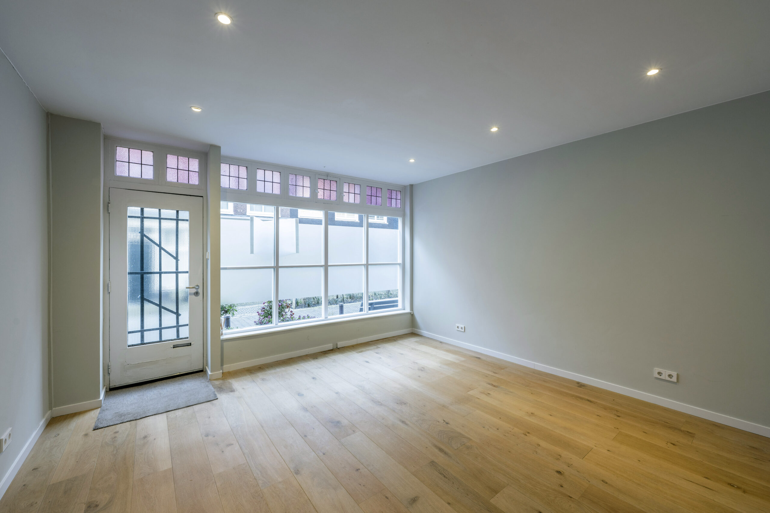 Lichte, lege woonkamer in de Leliestraat met houten vloer, grote ramen en een deur naar de voortuin.