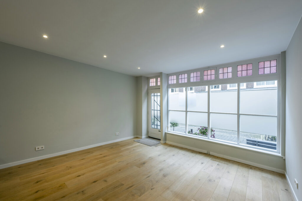Lichte, lege woonkamer in de Leliestraat met houten vloer en groot raam met glas-in-lood bovenlichten.