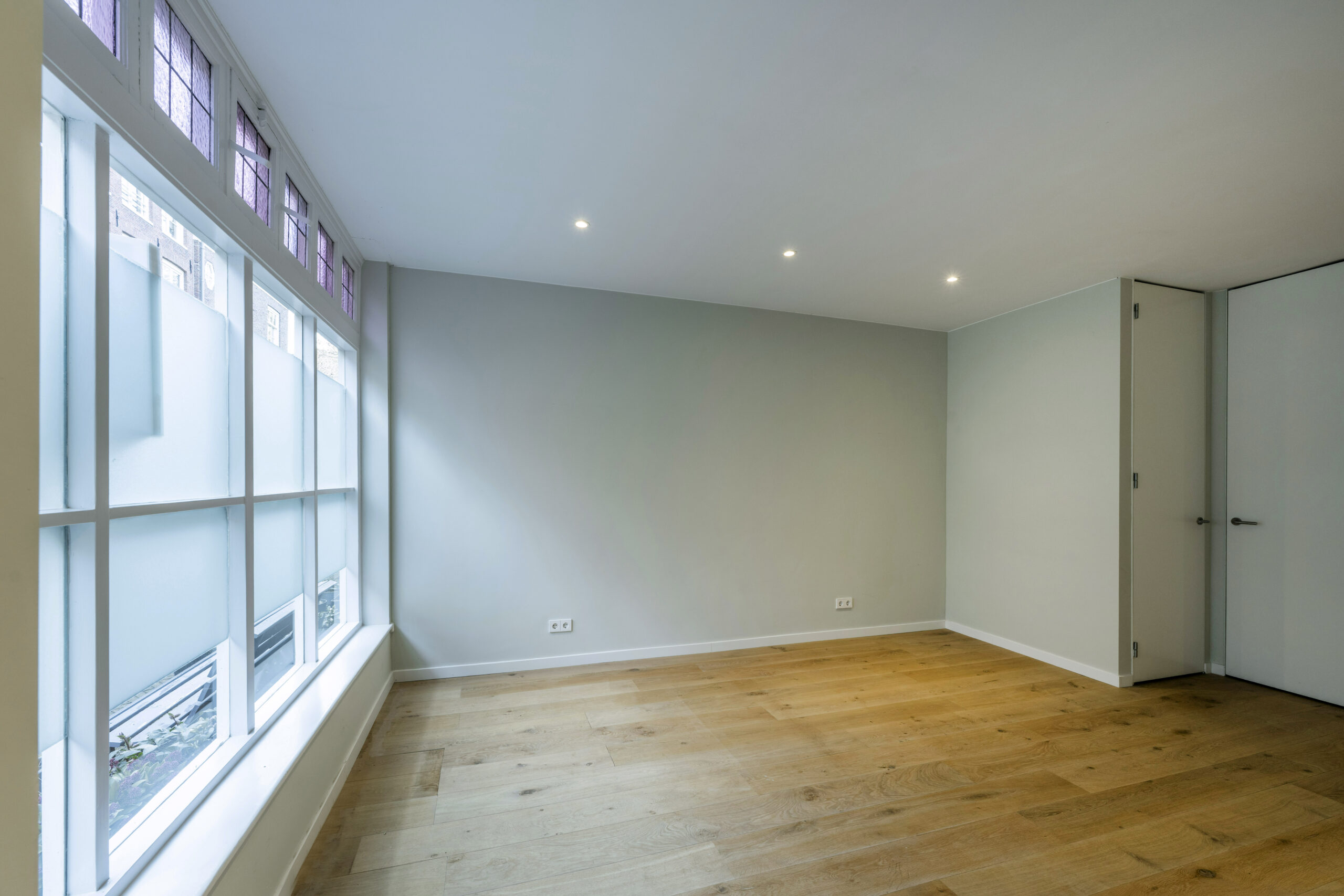 Licht en leeg vertrek met houten vloer, strakke witte muren en grote raampartij met glas-in-loodramen aan de Leliestraat 011, ontworpen door Paul Starink.