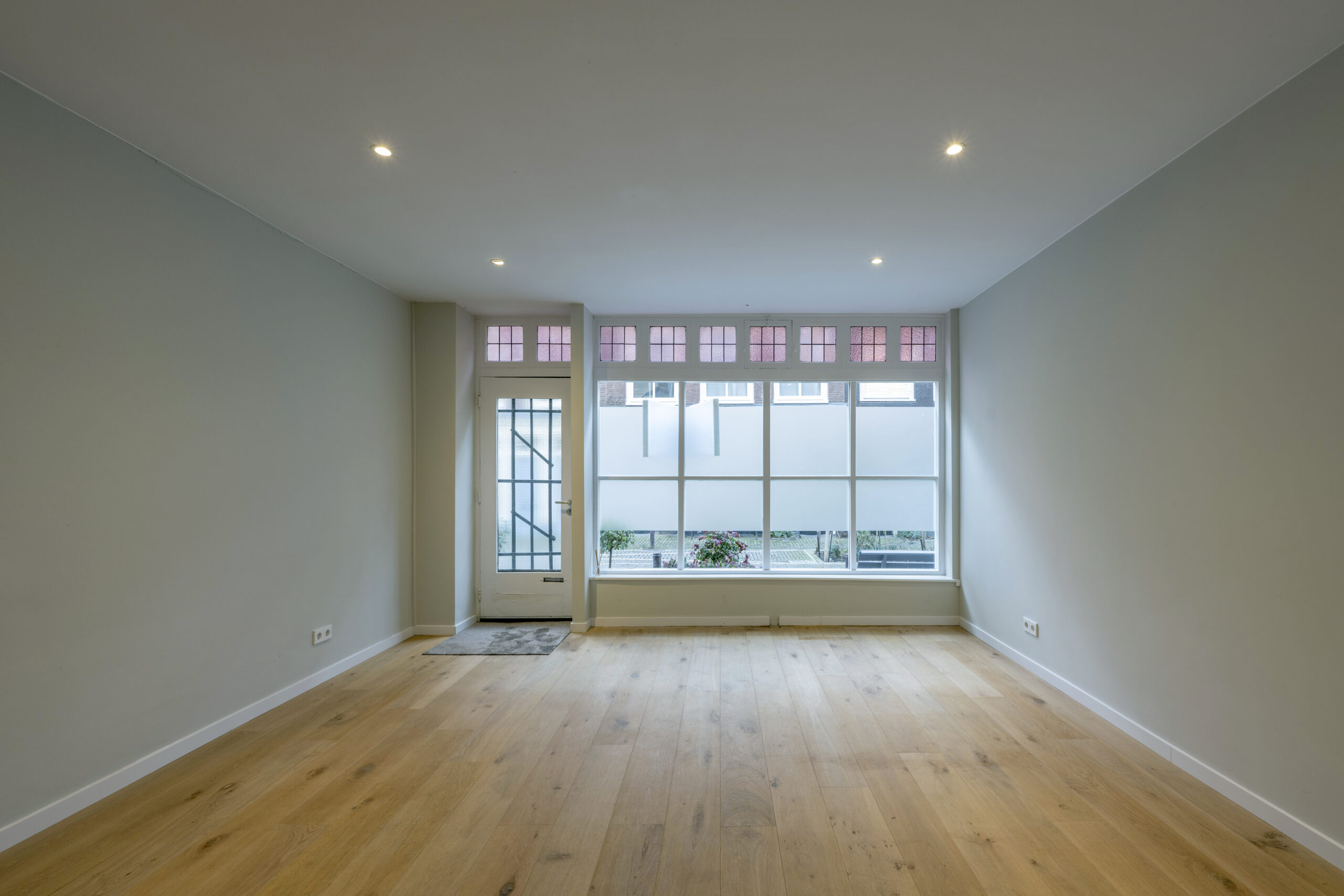 Lichte, lege woonkamer in de Leliestraat met houten vloer en grote ramen met glas-in-lood bovenlichten.