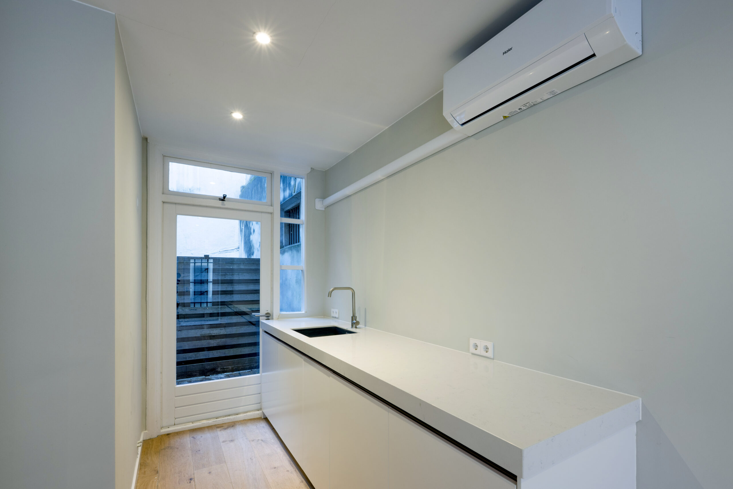 Strakke, moderne keuken in Leliestraat 016 met wit aanrechtblad, ingebouwde spoelbak en airconditioning aan de muur.