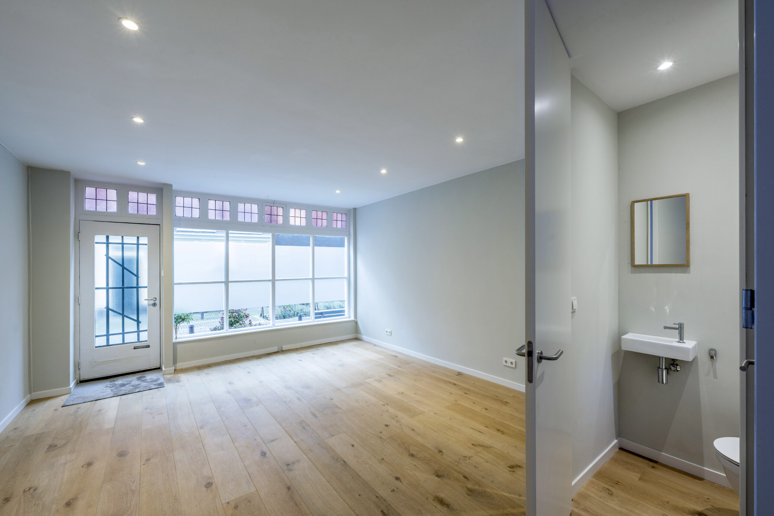 Lichte woonkamer aan de Leliestraat met houten vloer, grote ramen en aangrenzend toilet met wastafel.