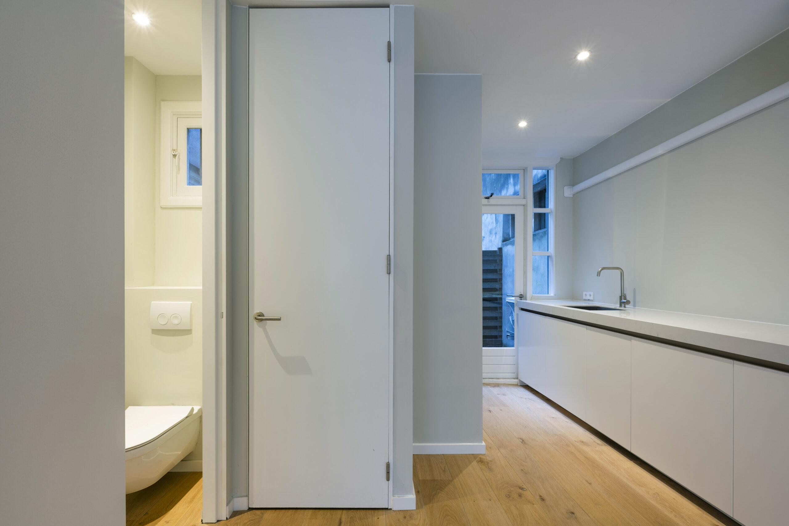 Moderne, minimalistische keuken aan de Leliestraat 020 met wit keukenblok, houten vloer en zicht op een toilet en achterdeur.