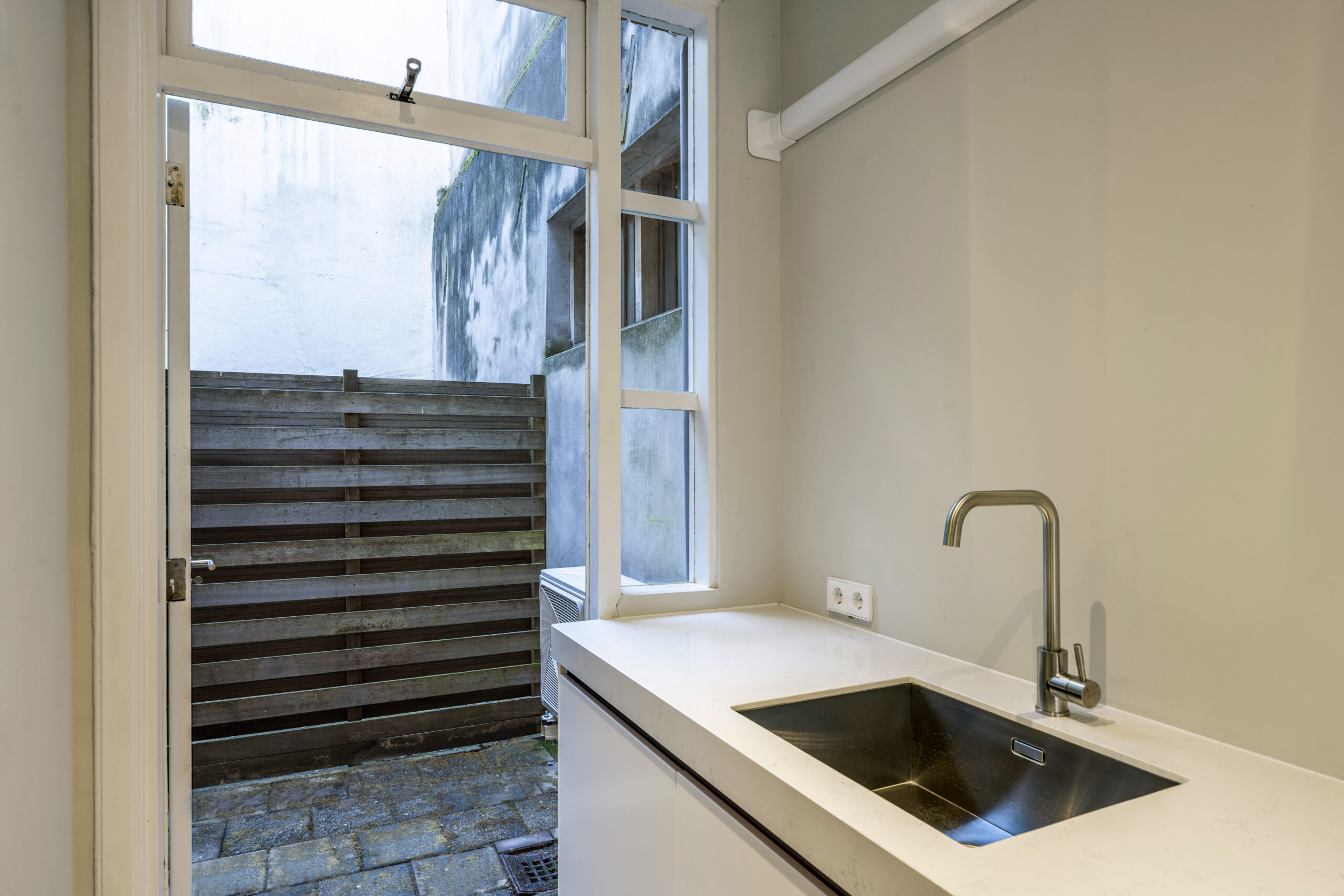 Keuken met wit aanrecht en rvs-kraan, grenzend aan een kleine betegelde buitenruimte met houten schutting, in woning aan Leliestraat 023 van Paul Starink.