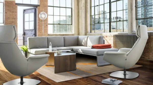 Moderne loungespace met grijze designmeubels, grote industriële ramen en een houten vloer.